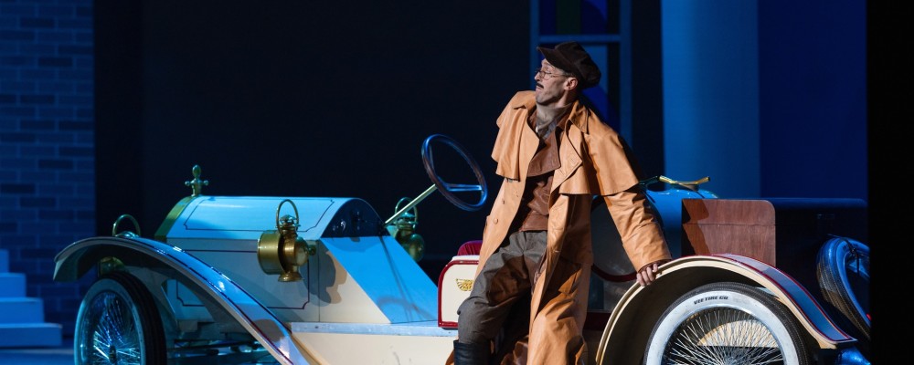Kadr z opery Don Giovanni, postać w brązowym płaszczu opierająca się o białe auto w stylu retro