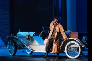 Kadr z opery Don Giovanni, postać w brązowym płaszczu opierająca się o białe auto w stylu retro