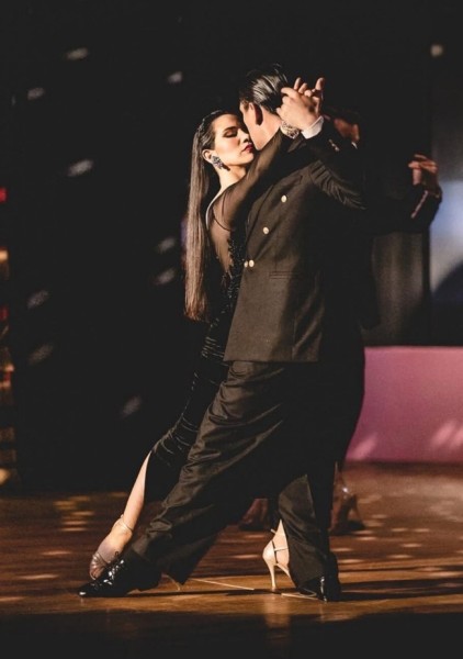 Na scenie para tancerzy w eleganckich, czarnych strojach wykonuje tango. Kobieta, ubrana w dopasowaną suknię z wysokim rozcięciem, z długimi, gładko rozpuszczonymi włosami, patrzy intensywnie na partnera. Jej twarz wyraża pełne skupienie i pasję. Mężczyzna w czarnym garniturze prowadzi zdecydowanym, eleganckim gestem. Oboje spleceni w bliskim objęciu tworzą pełną napięcia i emocji taneczną pozę. Drewniana podłoga sceny odbija światło reflektorów, a delikatne rozbłyski w tle podkreślają dramatyzm chwili.