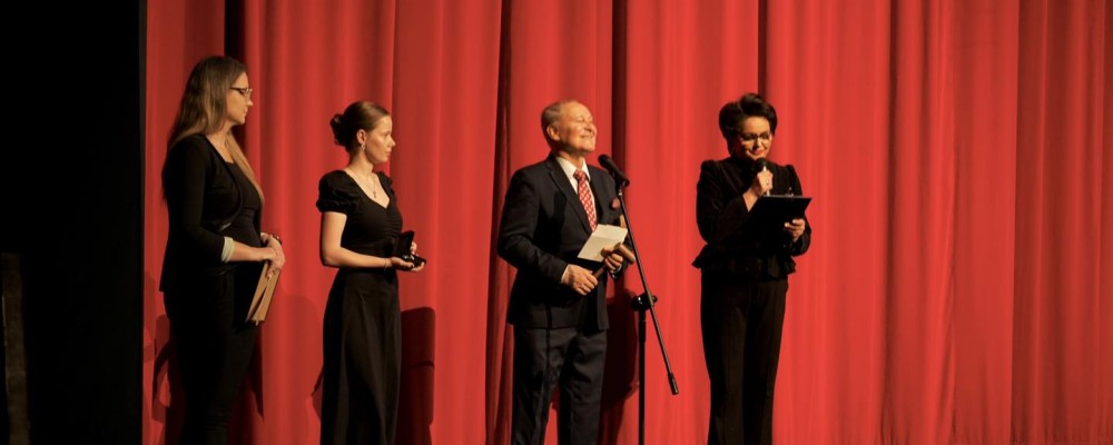 Czworo ludzi ubranych w czarne, eleganckie ubrania stoi na scenie prowadząc licytację charytatywną. W tle czerwona kurtyna.