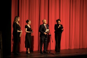 Czworo ludzi ubranych w czarne, eleganckie ubrania stoi na scenie prowadząc licytację charytatywną. W tle czerwona kurtyna.