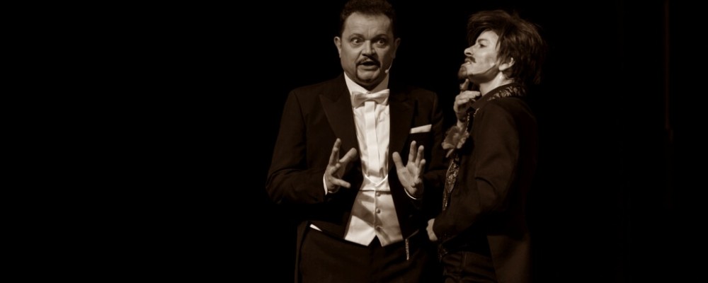 Janusz Ratajczak na scenie w eleganckim stroju gestykuluje obiema rękami. Obok stoi inny artysta. Zdjęcie jest w odcieniach sepii.