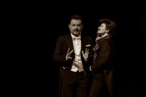 Janusz Ratajczak na scenie w eleganckim stroju gestykuluje obiema rękami. Obok stoi inny artysta. Zdjęcie jest w odcieniach sepii.