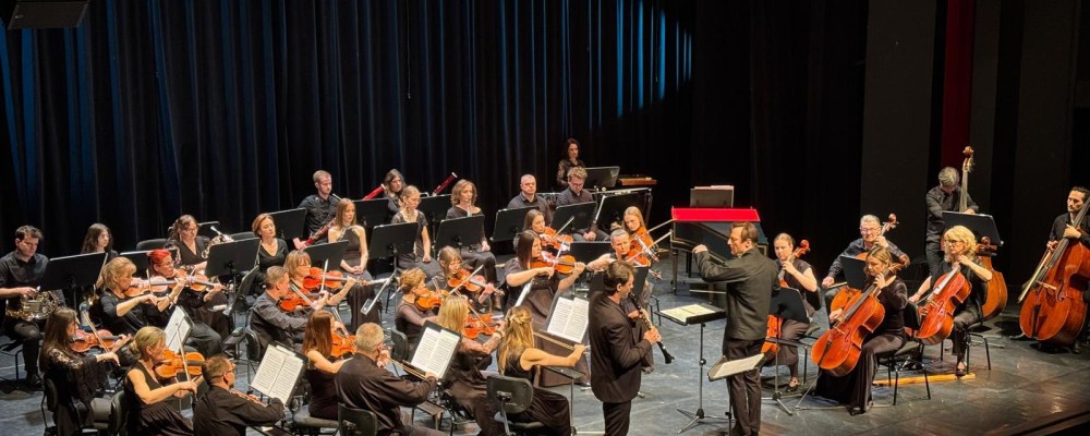 Na scenie występuje orkiestra z muzykami grającymi na instrumentach. Dyrygent stoi z przodu, twarzą do wykonawców. Scena jest oświetlona, a publiczność jest częściowo widoczna w prawym dolnym rogu.