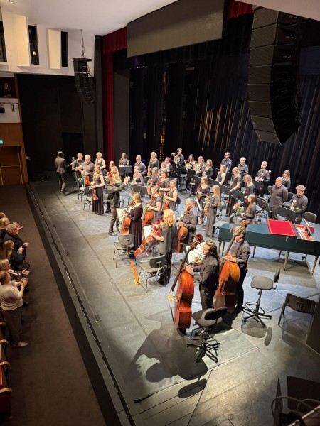 Muzycy orkiestry stoją na scenie trzymając swoje instrumenty, podczas gdy publiczność bije brawo. Dyrygent stoi z boku.