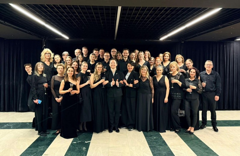 Zdjęcie grupowe stojących razem muzyków orkiestry ubranych w formalne czarne stroje.  Niektórzy trzymają w ręku kieliszki w tle widać czarne zasłony i jasne górne oświetlenie.
